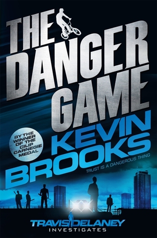 The Danger Game white letters. Kevin Brooks in blue letters. boy on bike beside The. boy and men behind him. blue sky and buildings behind him.