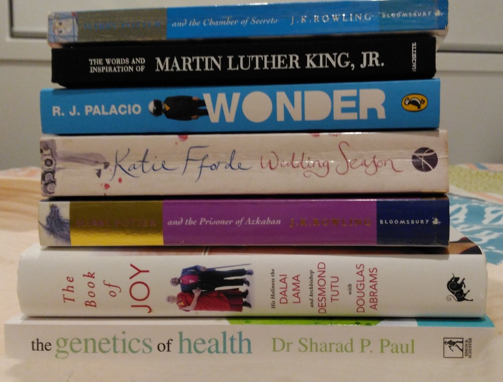 stack of six books on top of bed with window behind. colours of white, dark pink with gold, blue and black on spines of books. images of people and words along spine either horizontal or vertically spaced.