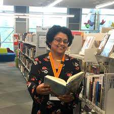 image of woman holding book inside library. lights above. woman in black and flowery dress with bright orange lanyard around neck.