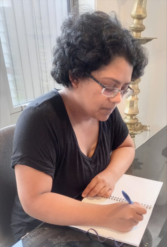 woman in black top seated at glass table writing in book with blue pen. window behind with golden lamp behind in corner.