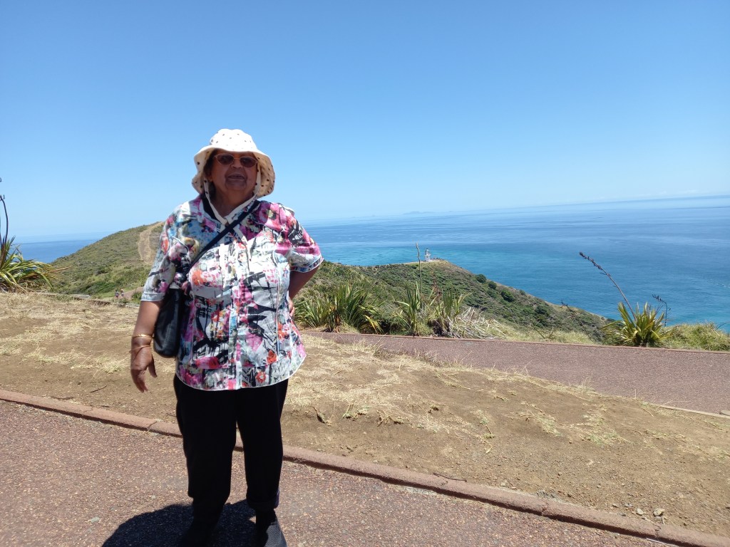 woman in hat with bag standing on track with view of sea and lighthouse on hill in back.