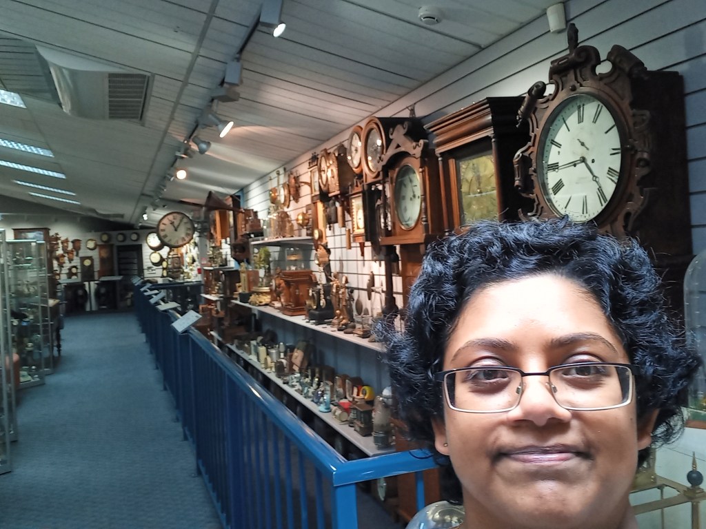 face of girl in glasses in right bottom. display of clocks of different sizes and shapes behind her in low-ceilinged building.
