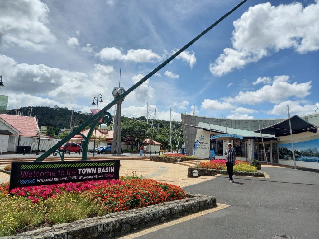 image of buildings in town centre. blue sky with clouds. sign beneath large pointy wand says Welcome to the Town Basin. man stands between flower beds and in front of building.