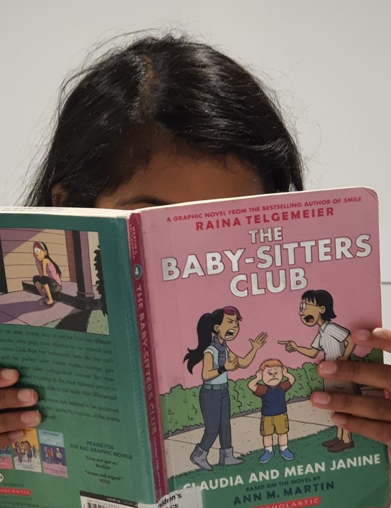 image of girl holding book. top of dark hair showing above pink and green The Baby-Sitter's Club book. pale grey background behind.
