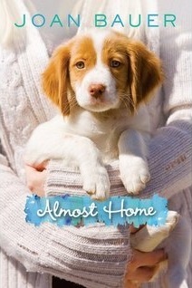 image of small white and brown dog held in arms of someone wearing white jumper. blue letters at top read Joan Bauer. white letters across blue in middle read Almost Home.