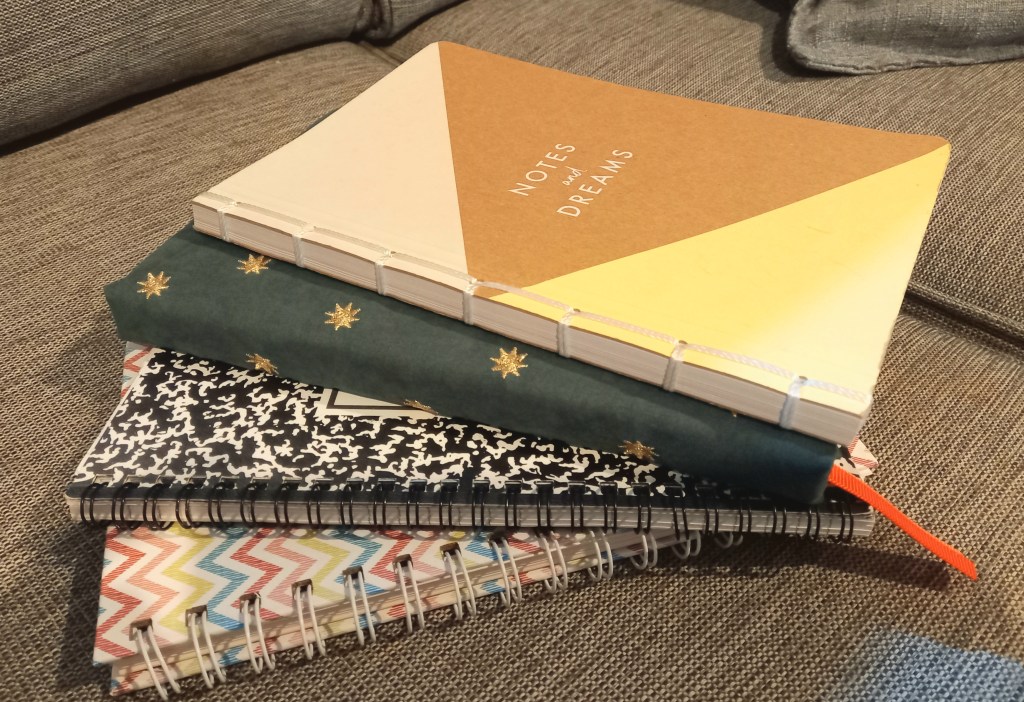 Small pile of four notebooks in spiral one on top of other. bottom two multi-colored and black and white. top two green with stars and pale colors on top with writing acorss middle reading Notes and Dreams. grey surface of cushions below them.