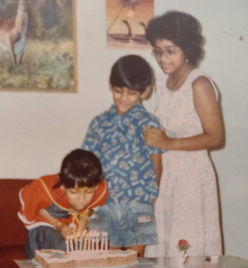 three kids in front of birthday cake. girl in red dress bending down to blow out candles. paintings of animal and boats on wall behind them.