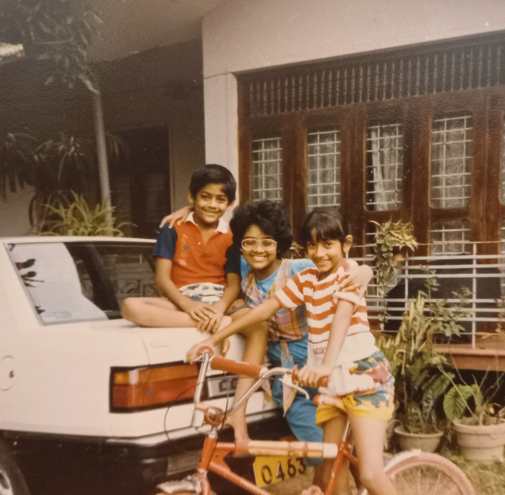 three kids outside house. girl in red striped tshirt and shorts on bicycle. boy in red shirt seated on top of back of car. girl with glasses in blue in between them. brown windows and cream walls behind them.