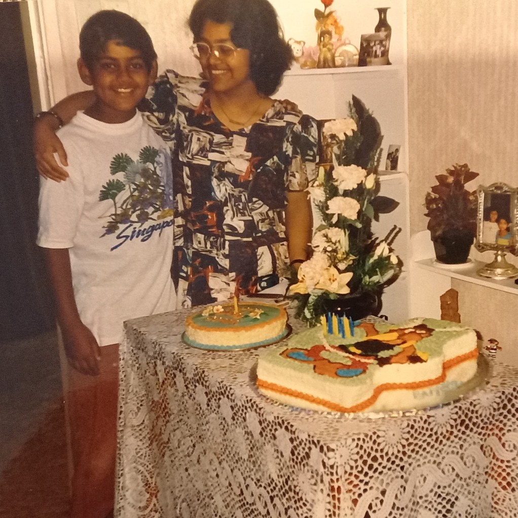 girl and boy standing in front of table with two birthday cakes. flower arrangement in between cakes and shelf behind them with trinkets. girl in colourful dress has arm across boy in white tshirt.