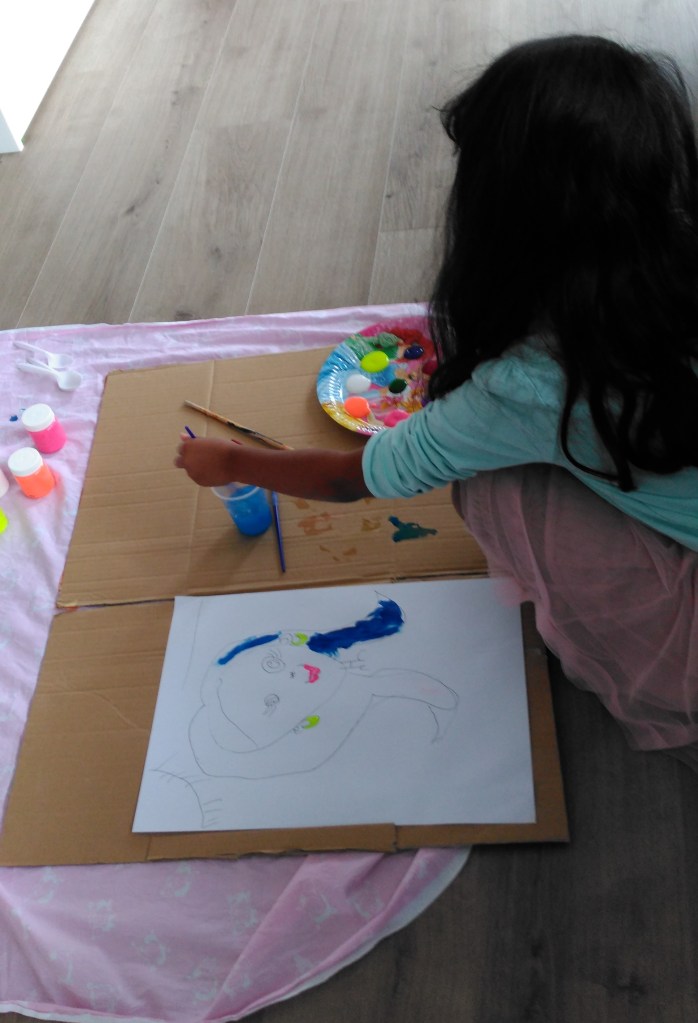 image of young girl with long dark hair in blue top and pink skirt painting on ground with paper and pots.