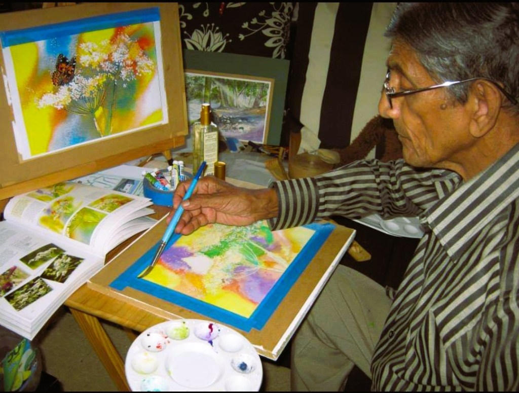 image of man with glasses in striped shirt painting on table with books and paints around him.