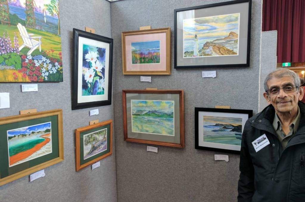 man in dark jacket standing next to grey screens with eight framed artworks mounted on them.