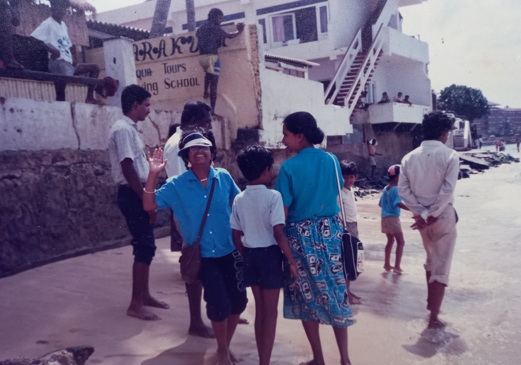 group of people along water edge. white building with steps. man on wall painting. woman in blue skirt and blouse with boy. girl in blue shirt and pants with white cap waving and laughing at camera.