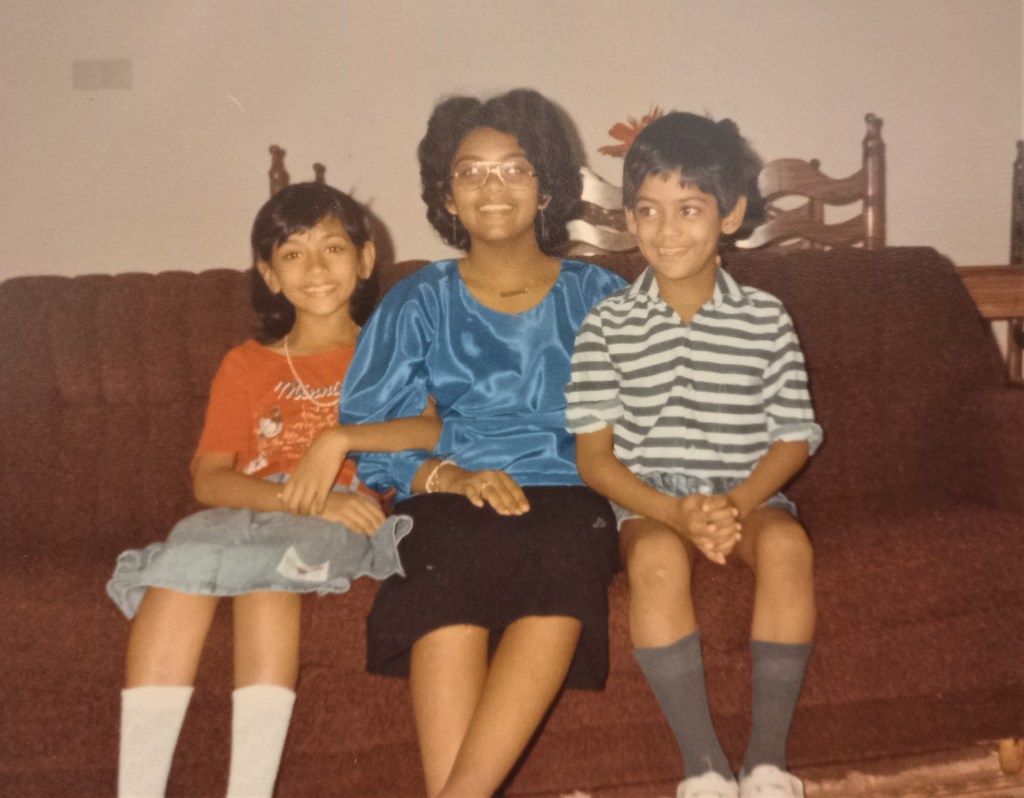 two girls and small boy seated on brown sofa with brown chair backs behind them. boy with striped shirt and shorts. older girl with blue blouse and black skirt and younger girl with red top and blue skirt.