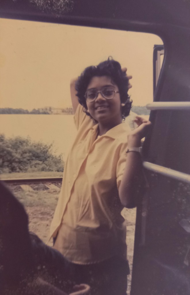 girl in yellow blouse posing beside open door of vehicle. lake behind her. fairly old quality to image.