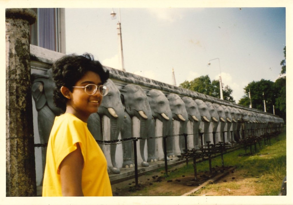 image of girl in glasses with yellow top posing in front of wall of elephant head sculptures on a sunny day.