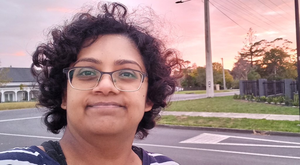 author outside with light pink sky. background has houses, road and fence.