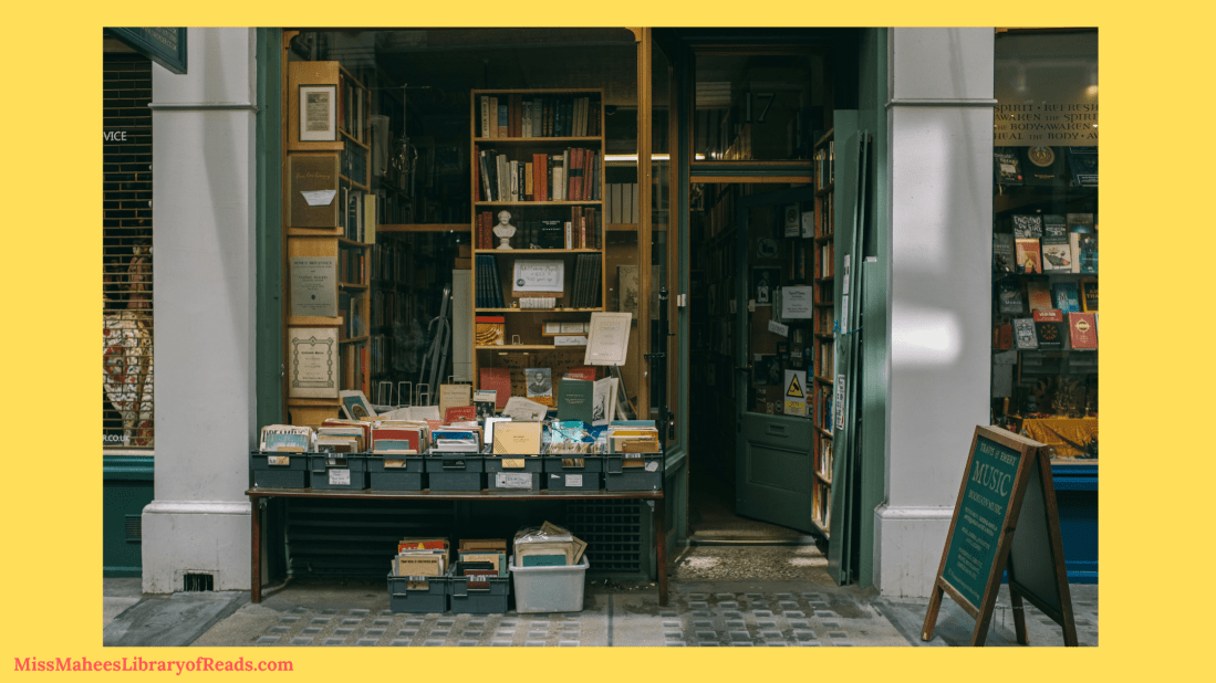 image of front of bookshop with shelving units filled with books inside. grey columns with large windows and green door. low table with green bins of books on it and on ground. green sandwich board in front of door. yellow background. red small letters on bottom left corner reads MissMaheesLibraryofReads.com.