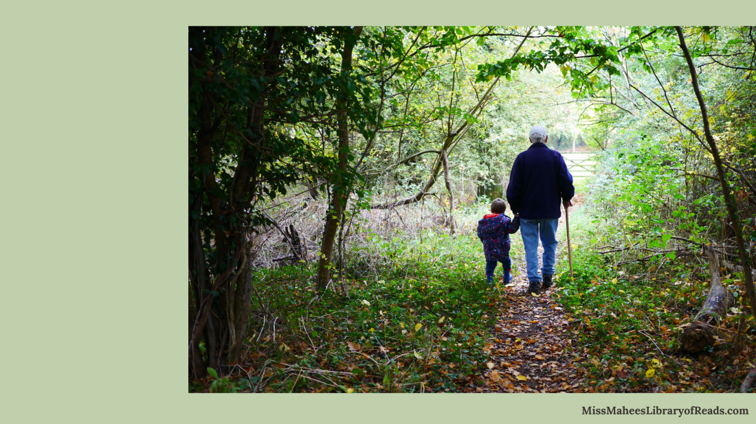 large image aligned right of elderly grandfather with walking stick in dark coat, jeans and white cap holding hands with small child in dark clothes walking along path in middle of forest like area with trees and grass and bushes on either side of them. light green background. MissMaheesLibraryofReads in small black letter at bottom right edge.